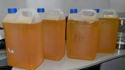 Large plastic containers are seen with orange THC liquid on a table
