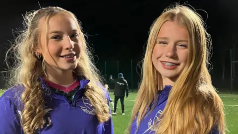 BBC Two young girls are smiling for the camera. They are both wearing purple zip up jumpers with their football club branding on them in white. They are standing on a football pitch which is lit up with flood lights. They both have long blonde hair and braces.