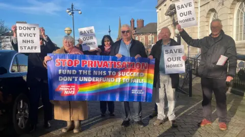 A small group of men and women hold up banners and posters outside an official looking building. The banners read: Save Our Homes