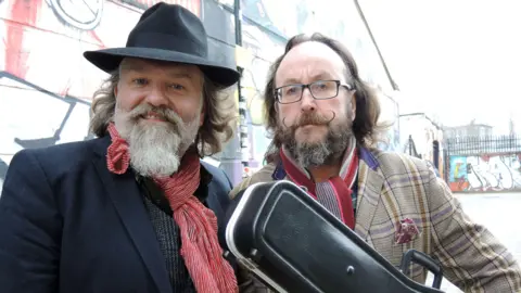 Si King and Dave Myers pose for the camera while filming one of their shows. King wears a black hat and Myers holds a guitar case.