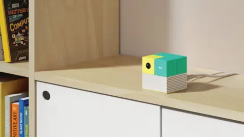 Nex Playground A close up on the Nex Playground console, a small green yellow and white cube on a surface, with white cables plugged into it, next to a bookshelf space