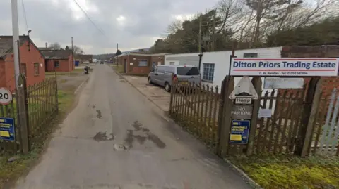 The entrance to an industrial estate with a road leading towards a number of single-storey brick buildings and a sign reading Ditton Priors Trading Estate