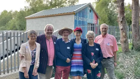 Tony Cowling Sally Waterman, Andy Tunstall, Tony Cowling, Sophie Fenwick-Paul, Anne Wheldon, David Whipple posing in front of Reading Hydro. Two turbine wheels can be seen in the distance with the house that says 'Reading Hydro'. 
