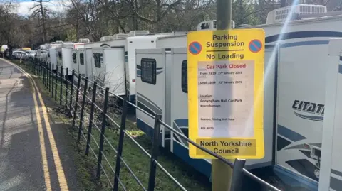 A yellow council notice on a lamppost details the car park closure. Several white lorries can be seen parked along the metal fence inside the car park.