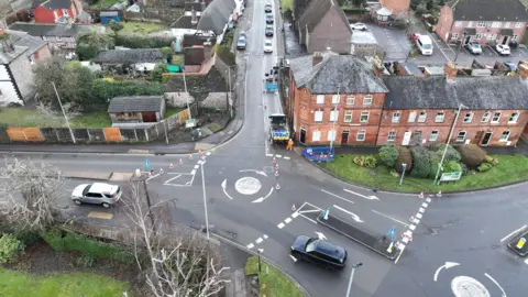 An aerial image of a roundabout surrounded by cones and blue scaffolding. In the distance, a line of cars wait.