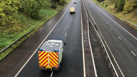 ATO team travelling along the motorway, one vehicle is clearly in shot with yellow and red stripes on its back panel, the roof is grey and the rest of the vehicle appears to be white.