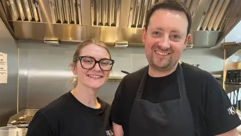 Nicola Haseler/BBC Two people dressed in black, smiling in a silver kitchen.
