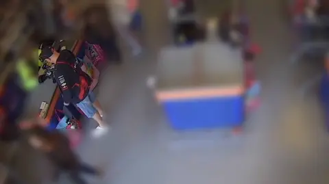 B&M Mr Long is seen at the till inside the B&M store paying for his shopping. The surrounding image is blurred. He is wearing a black and red jacket, blue jean shorts, a black cap and white trainers.