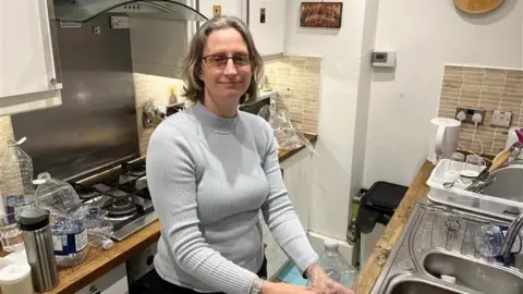 Phil Harrison/BBC A woman with light grey hair and wearing a grey polar neck sweater and black trousers stands in a modern kitchen.