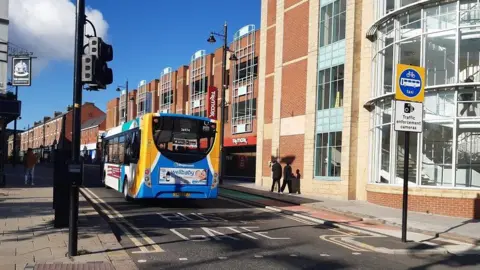 LDRS A single-decker bus driving down a street. 'Bus Gate' is written on the road and a sign with 'traffic enforcement camera' alongside a picture of a bike, bus and the word 'taxi'. Black cameras are pointing down the street.