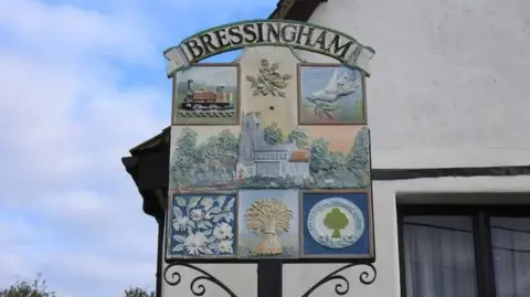 Adrian S Pye/Geograph village sign for Bressingham in Norfolk showing wheatsheafs, doves, trains and a church in the centre