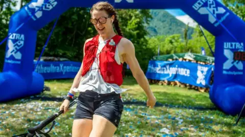 Gemma Hunter A woman cross the finish line at a race. She is wearing a white top and black camouflage shorts. She is also wearing a red running vest with a water bottle.She is also wearing sunglasses.