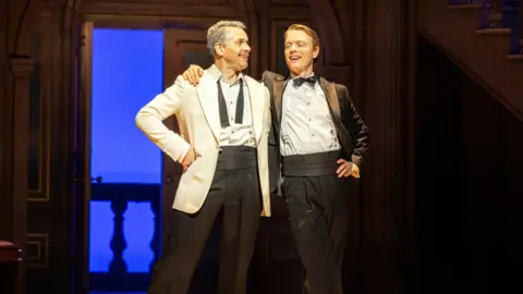 Trafalgar Entertainment Freddie Fox (right) wears a tuxedo and looks cheekily at his co-star in a white tuxedo jacket