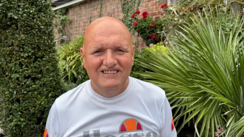 Martin Heath/BBC Rob with very short hair looking at the camera and wearing a white T-shirt with red and orange semi-circular logo. He is standing in front of ferns and trees in a garden.