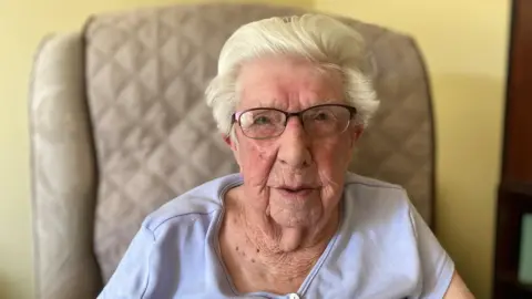 A woman with short white hair is looking at the camera. She is wearing rectangular metal glasses and a blue top. She is sitting in a chair.