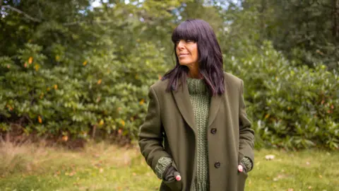 BBC/Studio Lambert/Llara Plaza Claudia Winkleman in a chunky green jumper with a dark green coat over the top. She stands in front of some bushes.