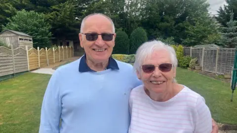 Mousumi Bakshi/BBC Roy and Sue Pettit in their garden. Roy is wearing a blue jumper and blue shirt, while Sue is where and pink hooped top. Both a smiling and looking at the camera