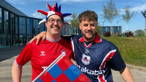 Two men stand side by side wearing York City shirts. The man on the left wears glasses, a blue, white and red jester-style hat and a red shirt. The man on the right has short brown hair and a beard, and a blue shirt.