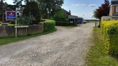 The access road into Folly Farm. It is a gravel road leading to a car park and farm gate in the distance. On either side of the road, there are houses, and on the left is a sign with "Tudor Place" written on it.