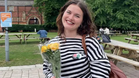 A young woman with long dark hair smiles at the camera and holds a yellow bunch of flowers. She is wearing a black and white striped top. Behind her is picnic benches and green space. 