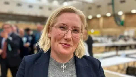 Labour MP Emma Lewell at the South Tyneside election count. She has shoulder-length blonde hair and wears glasses. She is wearing a necklace with a crucifix around her neck.