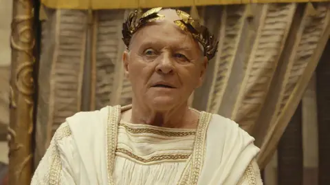 Peacock/Prime Anthony Hopkins dressed in white and gold emperor clothes stands in front of a curtain with soldiers to his left