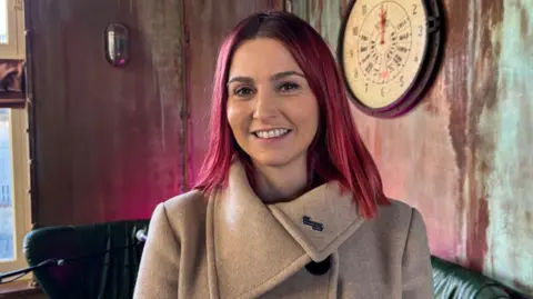 A woman with red shoulder-length hair is wearing a beige coat and smiling at the camera. Behind her is a large round clock on the wall and dark leather seats against a fading red wall.