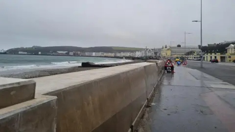 The thick, beige concrete wall, you can see the war memorial, villa marina and Fouglas head in the background, with construction taking place.