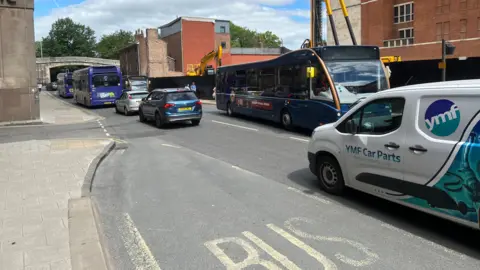 A picture of Rougier Street in York with cars and buses queuing in each direction. A demolition site, with yellow plant machinery, is to the right of the image. York's Bar Walls can be seen at the top of the picture.