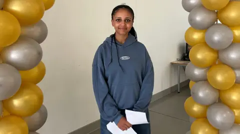 A teenager wearing a hoody stands under a balloon arch with results in hand.