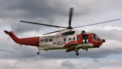 Irish coastguard helicopter in the sky. It is red and white. There are grey clouds in the background. 