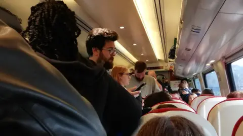 A view of an overcrowded CrossCountry train. A lot of people are standing on the isle. All seats are taken