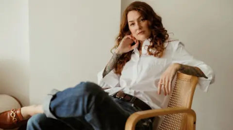 Olivia Steuer Maisie is sat on a wicker chair in a room painted white. She is wearing a white shirt with jeans and brown shoes. She has long red hair and tattooed arms.