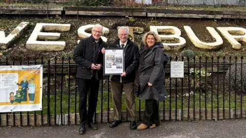 Horace Pickett posing for a picture standing beside two people outside the Westbury train station. He is holding an A3 frame in his hands, showing photos from his time on the team.