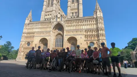A group of cyclists in cycling gear with bikes stood in front of Lincoln Minster. The sky is blue.