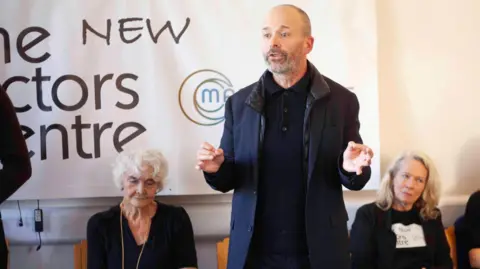 Gavin Watson Man in dark clothing addresses audience in front of a sign for The New Actors Centre at Marylebone Theatre