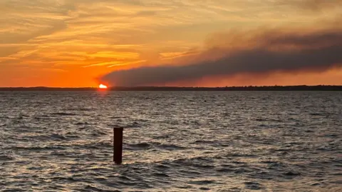 nomolos Looking across the sea to the coast in the distance. The sun is setting. A big dark plume of smoke is travelling across the sky.