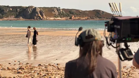 A camera man is seen from behind filminf two women walking along a sandy beach with cliffs in the background. 