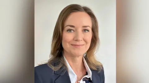 Elizabeth Stephens smiling with shoulder length brown hair, a blue suit jacket and a white blouse. She has a necklace with a pearl pendant. There is a plain beige background.