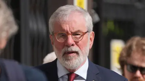 Gerry Adams is wearing orange round glasses and has short white/grey hair and a white/grey beard. He is wearing a navy suit jacket, red tie and a light blue shirt. He has two pins on his lapel, one is a gold circle and the other a red flower on a white background. 