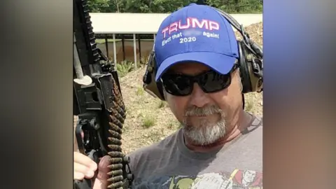 Facebook Johnny "Taz" Mulford, a man with a greying goatee beard, wearing a Trump 2020 hat, sunglasses, ear defenders and a comic book T-shirt, holding a gun with an ammunition belt hanging from it