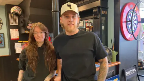 Chloe Millington and Jay from Barbertown in Droitwich. Millington is wearing a black hoodie. She has long brown hair and is wearing glasses. Jay is wearing a beige cap and a black top.