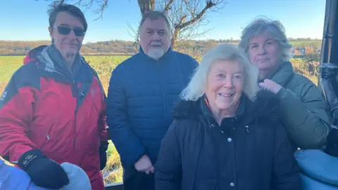 Amy Holmes/BBC A picture of four people standing outside with a field behind them. On the left of the image are two men, on the right two women. The man on the far left wears sunglasses, a red raincoat with thick gloves. The man next to him has a white beard and moustache and is wearing a blue coat. The woman on the far right is wearing a light green coat. Standing in front of her is a woman with slightly longer white hair and a dark blue coat.