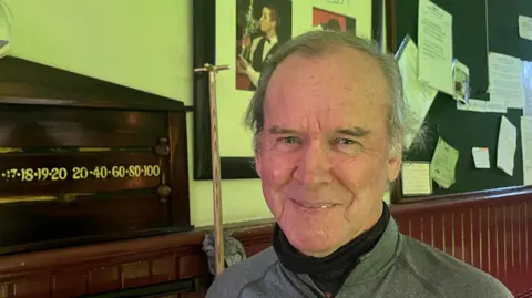 Andrew Turner/BBC David Stephenson, wearing a grey top over a black polar neck. He is standing next to a snooker scoreboard. 