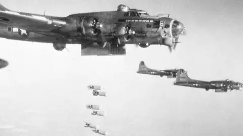 Getty Images A black and white picture of several Flying Fortresses flying in formation over land. They are four-engine monoplanes. A line of bombs has emerged from beneath one of them.