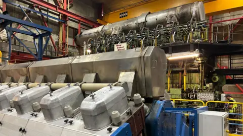Metal electricity equipment is shown within a concrete structure. Yellow, red and blue pipping and structures line the walls.