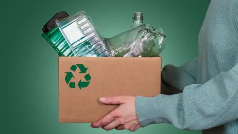 A person in a green jumper, pictured against a green background, holding a brown cardboard box that has a green recycling logo on it. The box contains plastic waste - bottles and food trays.