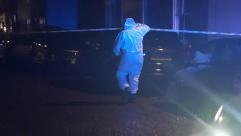 Joe Weir/ BBC A person in white overalls and black boots lifting and ducking under a blue and white police cordon. There are cars parked in a line on the right of the street. The night is dark.