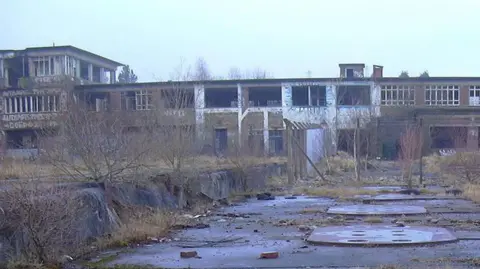 Robert Wade/Geograph The deserted site of Huncoat power station. It is a long two-storey brick building with a taller section to the left which have been vandalised. To the front are overgrown flat areas with covers on the ground and sprouting weeds