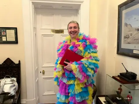 Joe Lycett stands outside the Lord Mayor of Birmingham's office door. He is wearing a layered, tulle style outfit, which is yellow, blue and pink.